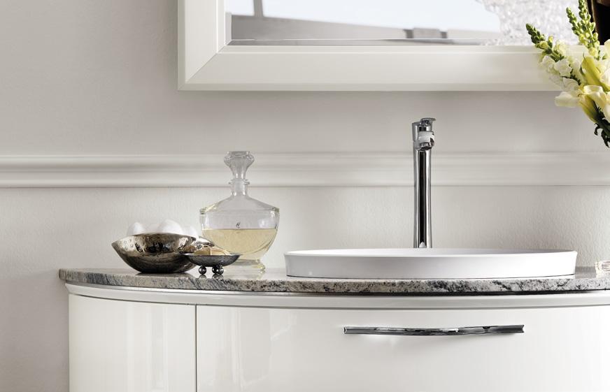 A modern bathroom sink with a sleek faucet, set on a marble countertop. Decorative items including a glass decanter and a metallic bowl are beside the sink. Flowers in a vase are partially visible on the right.