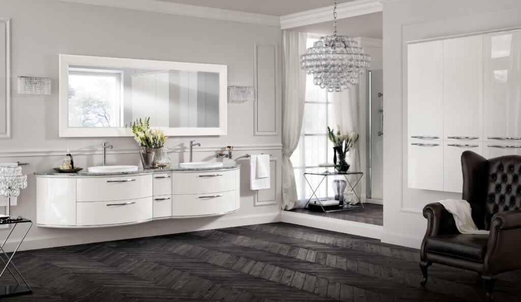 Modern bathroom with double sinks, white cabinets, a wall mirror, chandelier, dark herringbone floor, and a leather armchair.