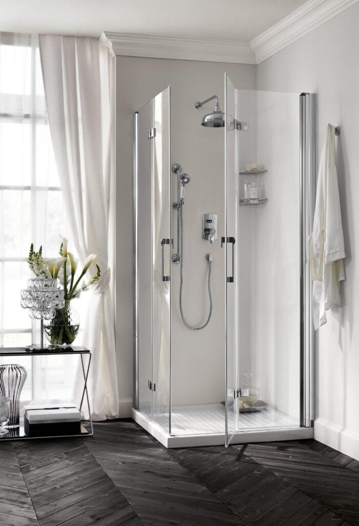 Modern shower enclosure with glass doors in a bathroom with dark herringbone wood flooring, a small table with a decorative lamp and flowers, and a robe hanging on the wall.