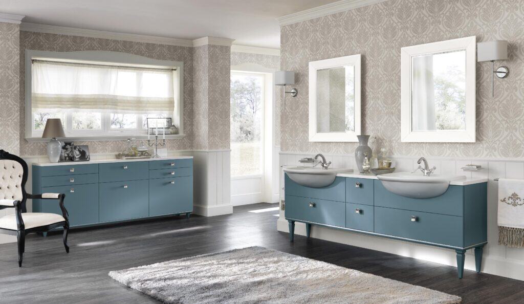 Spacious bathroom with blue cabinets, two white sinks, large mirrors, ornate wallpaper, and a plush area rug on a dark wood floor.