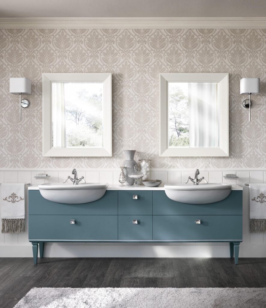 A modern bathroom with two sinks mounted in a teal vanity, featuring decorative mirrors, patterned wallpaper, wall sconces, and a gray area rug.
