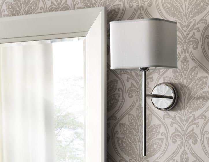 Wall-mounted lamp with a white shade on patterned wallpaper next to a window.