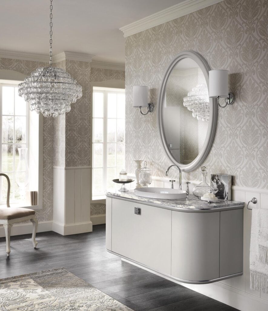Elegant bathroom with a wall-mounted vanity, marble countertop, oval mirror, and a crystal chandelier, complemented by patterned wallpaper and dark wood flooring.