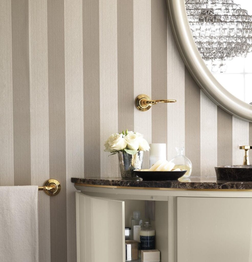 Elegant bathroom with striped wallpaper, round mirror, marble countertop, brass fixtures, white roses in a silver vase, and a towel on a gold holder.