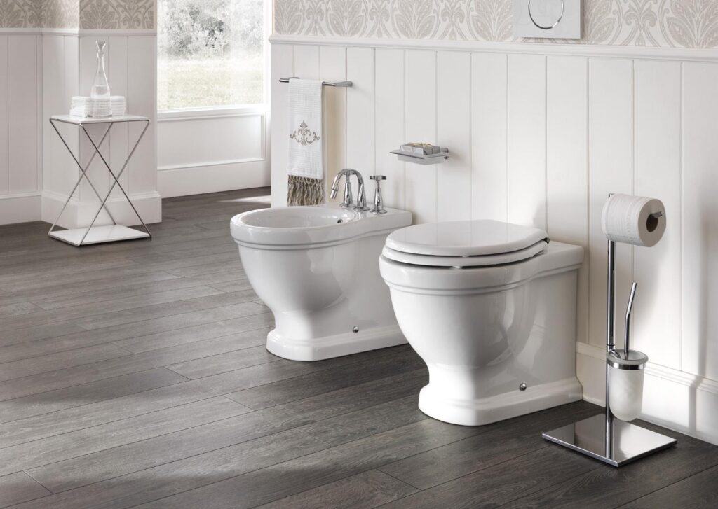 Bathroom with a toilet and bidet on wooden flooring, featuring a toilet-paper holder and a small side table with towels and a carafe.