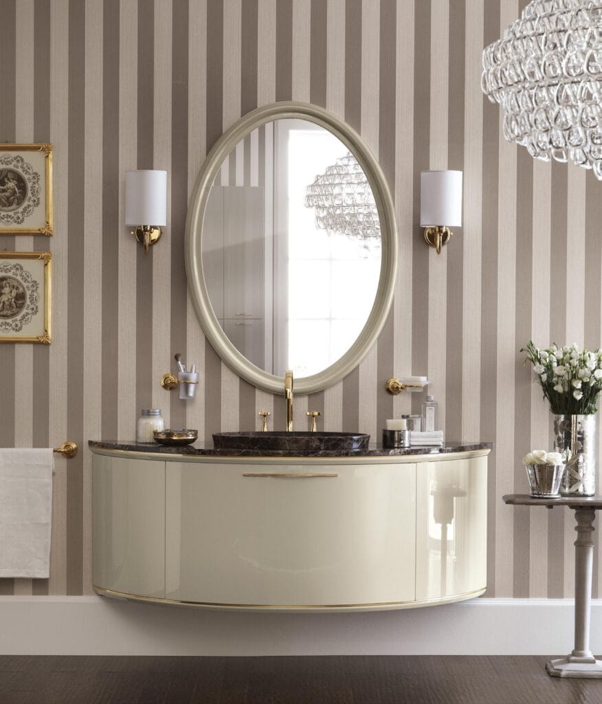 Elegant bathroom with an oval mirror, striped wallpaper, wall-mounted countertop with dark marble, gold fixtures, and a polished chandelier.