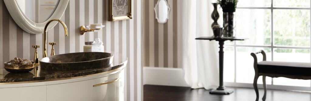 Elegant bathroom with a marble sink and brass fixtures against striped wallpaper, featuring a large mirror and a side table near a window with sheer curtains.