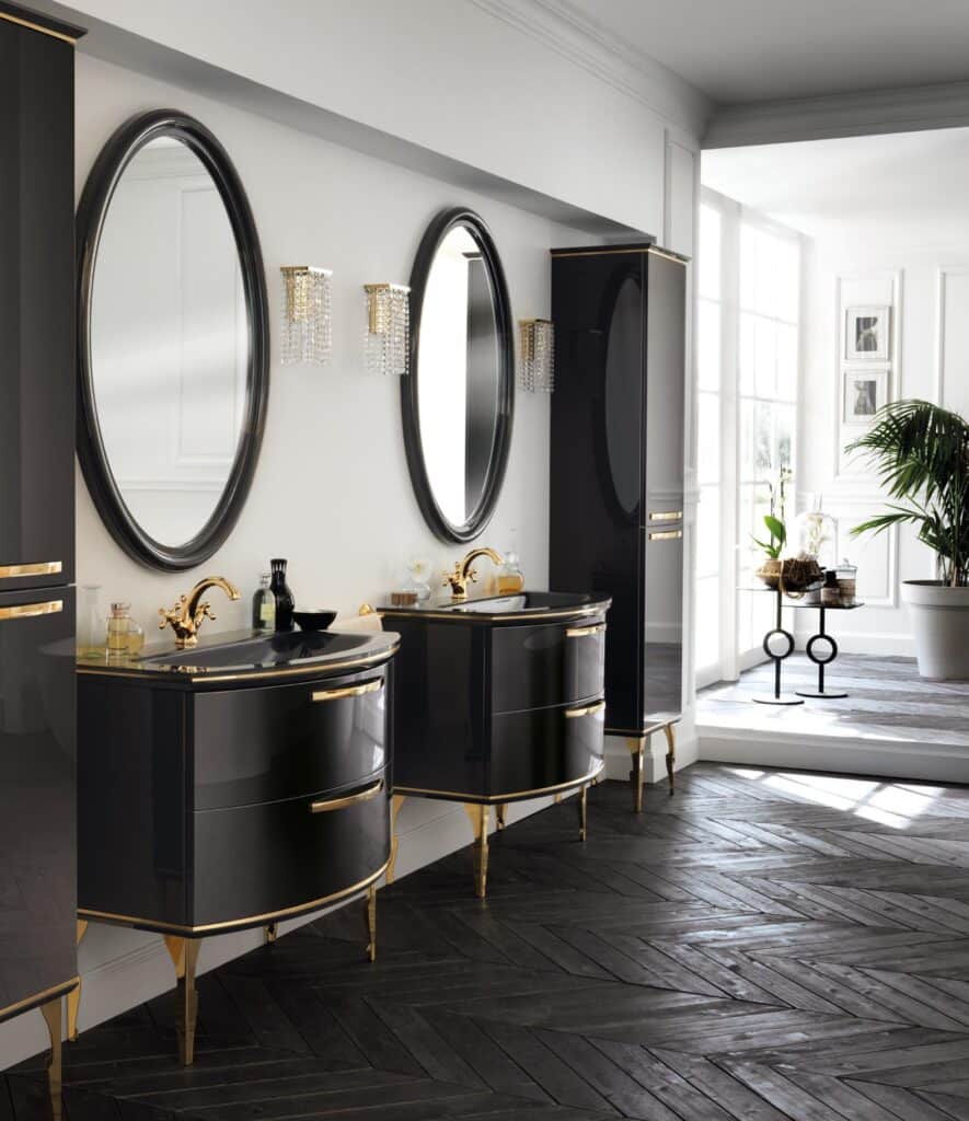 Luxurious bathroom with two black vanities featuring gold accents, oval mirrors above each vanity, crystal wall sconces, dark herringbone wood flooring, and large windows with natural light.