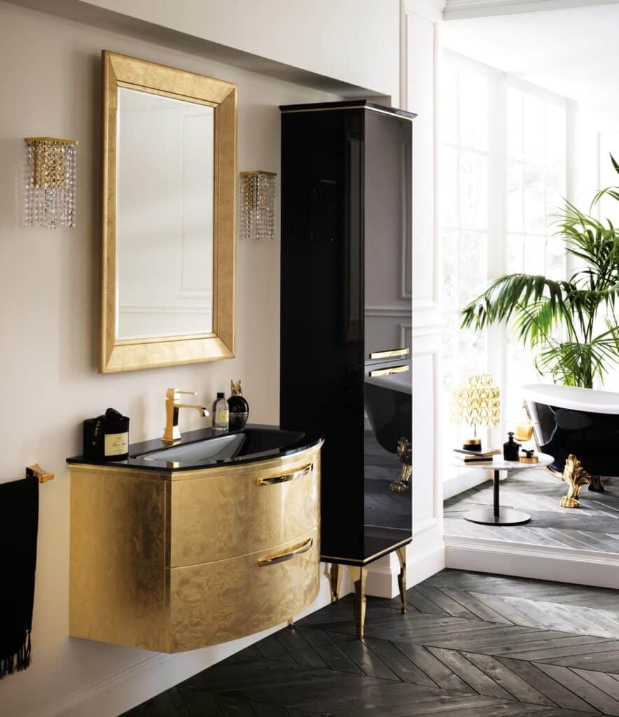 Luxurious bathroom with gold and black vanity, gold-framed mirror, crystal wall sconces, dark herringbone floor, and a bathtub with claw feet.