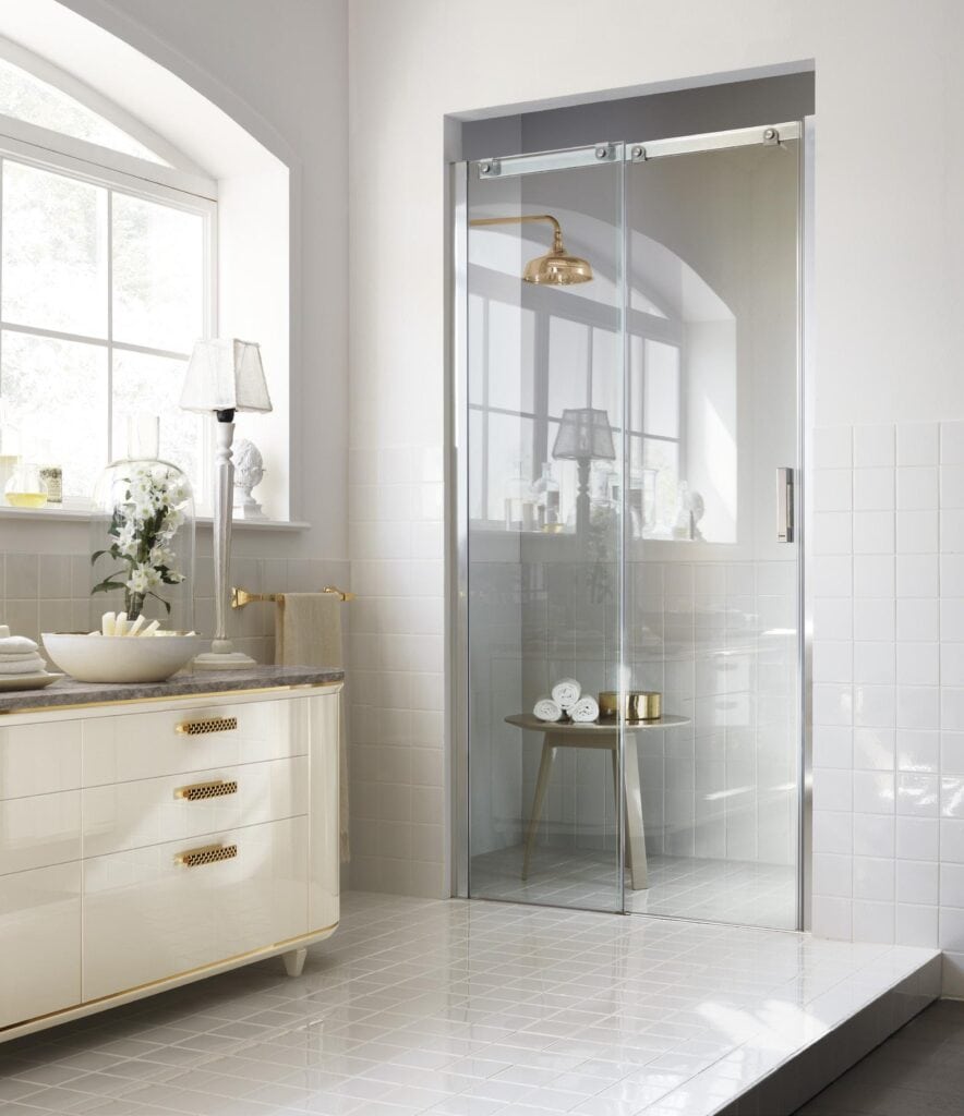 Elegant bathroom with a glass shower door, white tiled walls, and floor, featuring a vanity with gold accents and a small side table with rolled towels.
