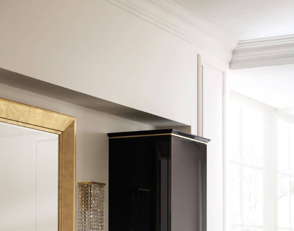 A partial view of a room with a gilded frame mirror, a black furniture piece, and rectangular wall lights, with large windows in the background.
