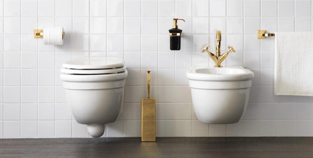 White tiled bathroom with a wall-mounted toilet and bidet, gold fixtures, toilet paper holder, towel rack, and a soap dispenser.