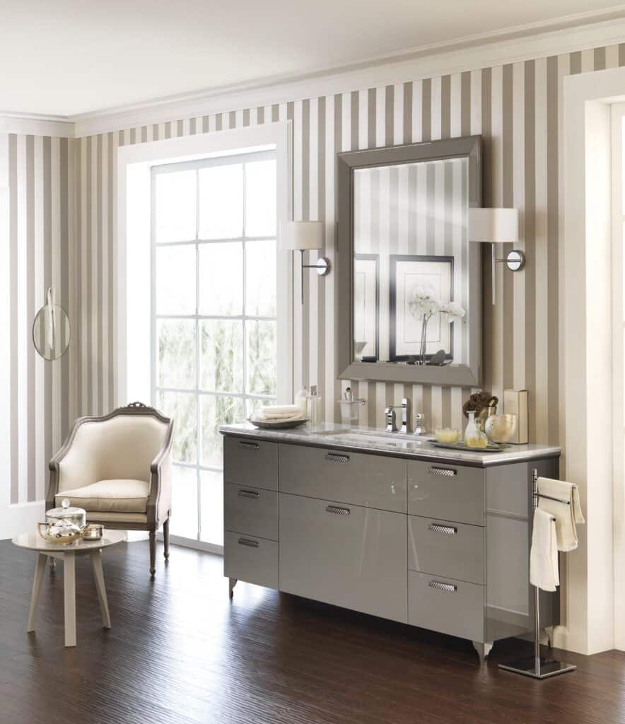 Elegant bathroom with a striped wallpaper, large mirror, wall sconces, a modern sink vanity, an upholstered chair, a small side table, and a window with natural light.