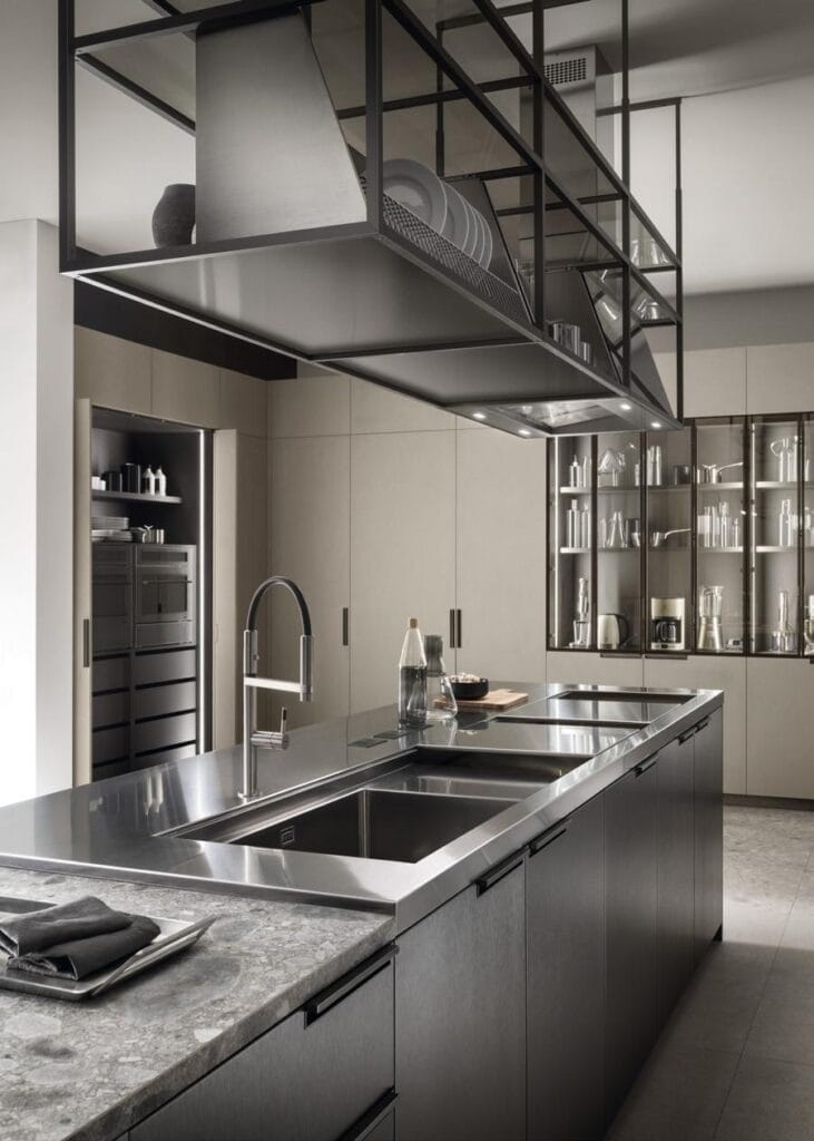 Modern kitchen with stainless steel appliances and countertops, featuring a large island with a sink and overhead shelving.