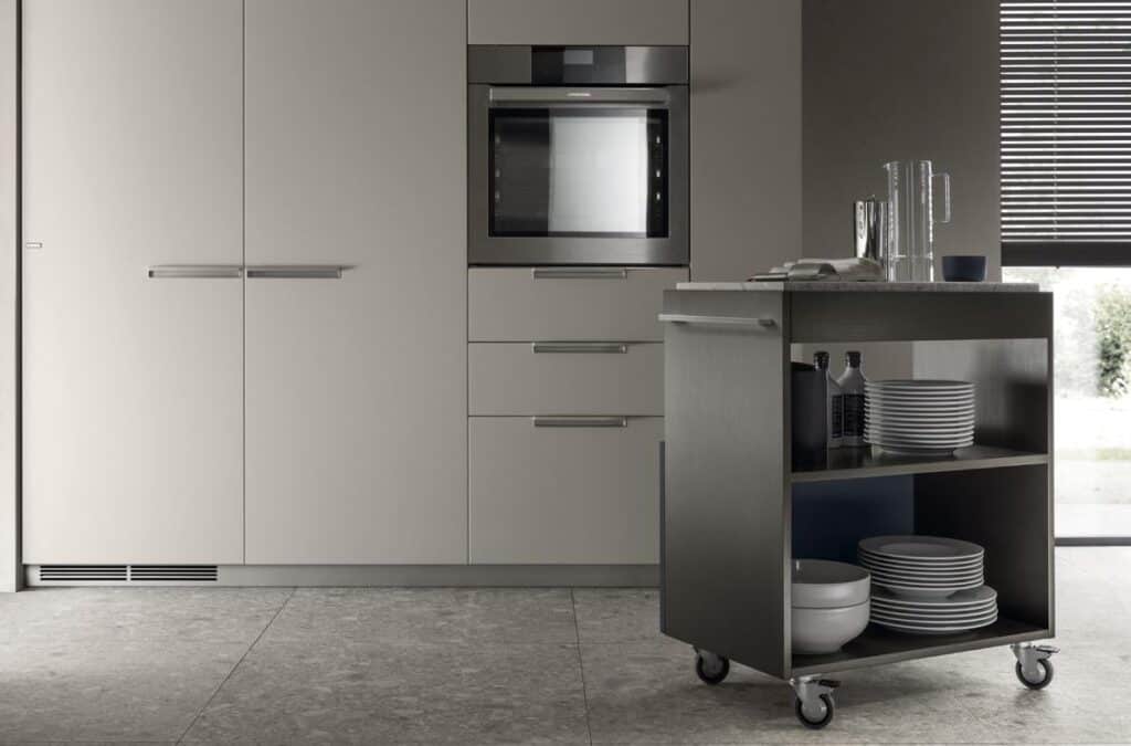 Modern kitchen with integrated oven, minimalist cabinets, and a mobile cart with shelves holding dishes and bottles.