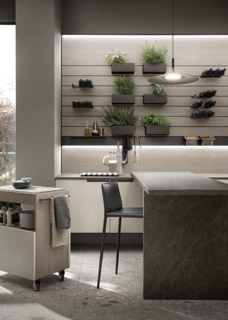 Modern kitchen with a dark stone island, black chair, wall-mounted plant holders, wine racks, and a pendant light.