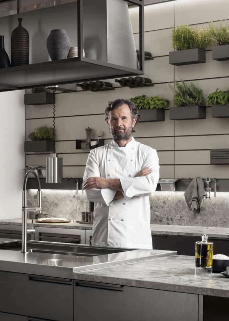 Chef standing in a modern kitchen with arms crossed, surrounded by herbs and kitchenware.