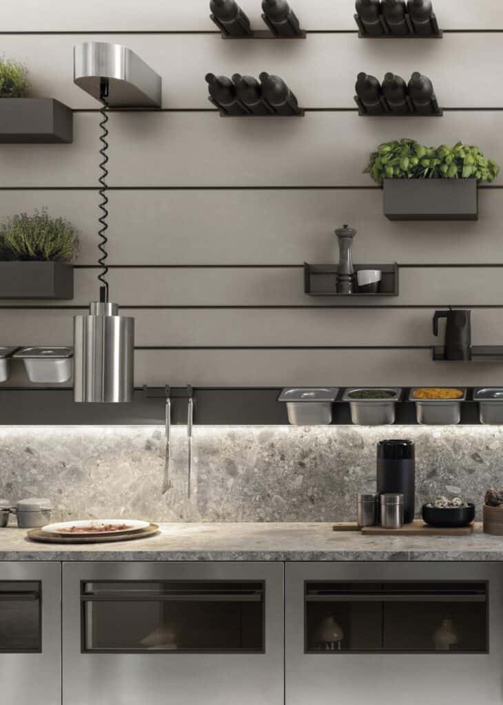 Modern kitchen interior with stainless steel pendant light, herb planters, shelves with bottles, spices, and a countertop featuring containers and utensils against a stone backsplash.