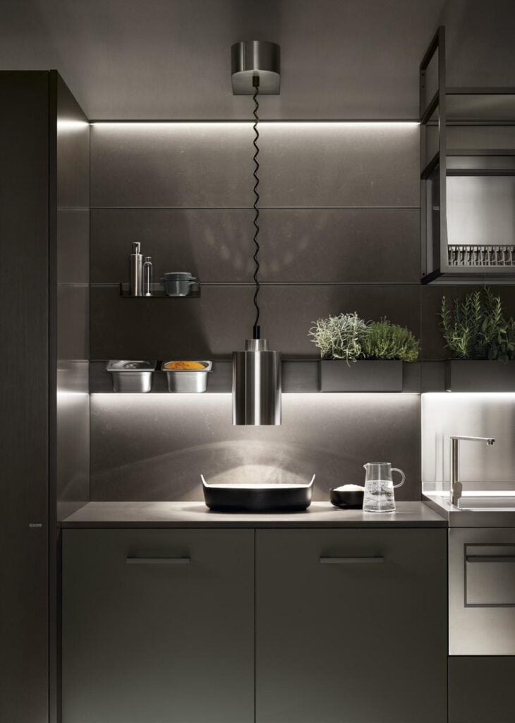 Modern kitchen interior with sleek cabinetry, a pendant light, potted herbs on a shelf, and a countertop holding a pitcher and bowl.