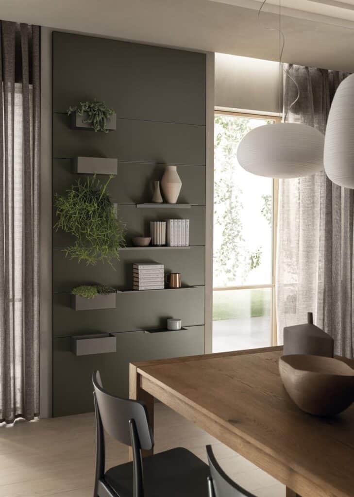 Modern interior with a wooden dining table, black chair, and wall-mounted shelves displaying books, plants, and vases; large windows with sheer curtains and hanging pendant lights.
