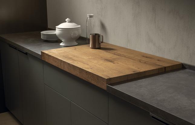 Wooden countertop extension on a kitchen counter, with a white soup tureen, plates, copper pitcher, and a glass bottle.