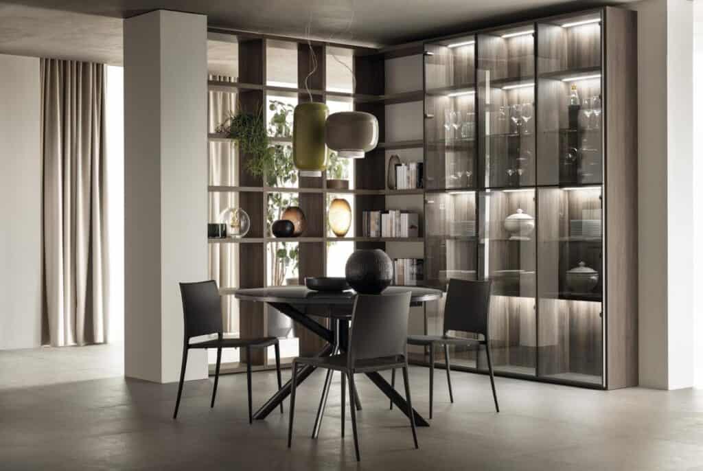 Modern dining room with a round table, four chairs, glass cabinet with dishware, open shelving with books and decor, and pendant lights.