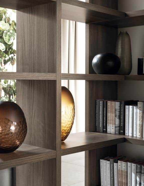 Wooden bookshelf with books and decorative vases placed on the shelves, with a glimpse of a plant in the background.
