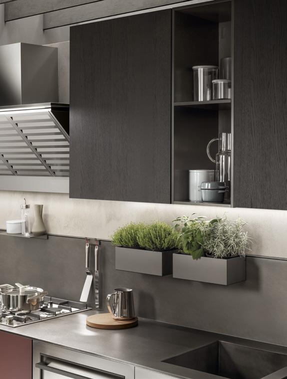Modern kitchen with dark cabinetry, metallic backsplash, open shelves, potted herbs, stovetop with pots, and a sink.