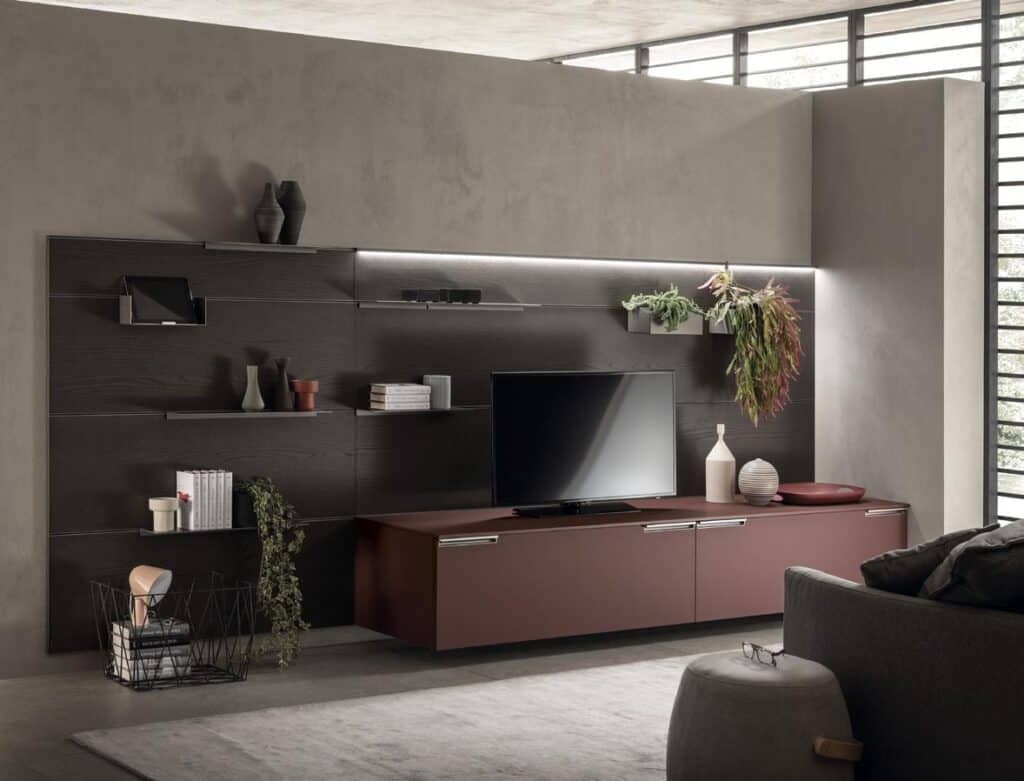 Modern living room with a TV on a sleek brown cabinet, wall-mounted shelves with decorative items, plants, a basket with magazines, and a gray sofa.