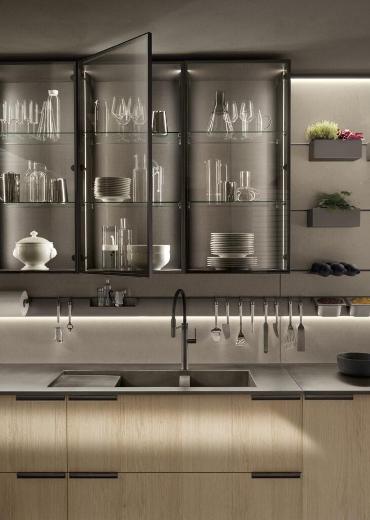 Modern kitchen with glass-front cabinets showing glassware and dishes, a sleek sink with a high-arc faucet, and wall shelves with plants and kitchen tools.