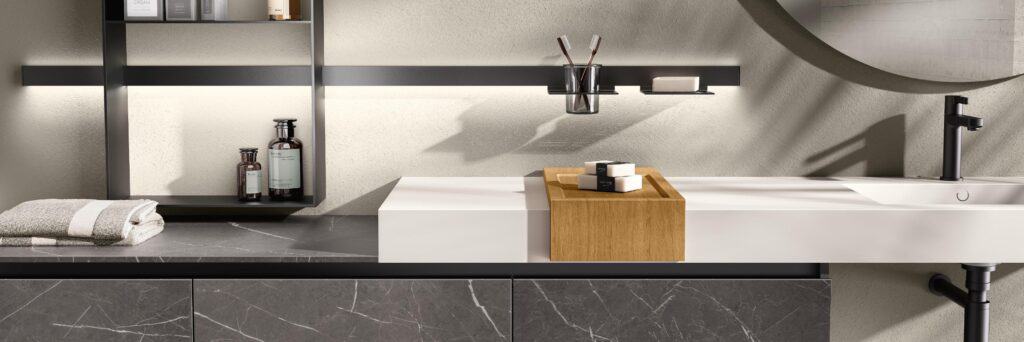 Modern bathroom vanity with a black faucet, white countertop, wooden tray, and wall-mounted shelves holding toiletries and towels.