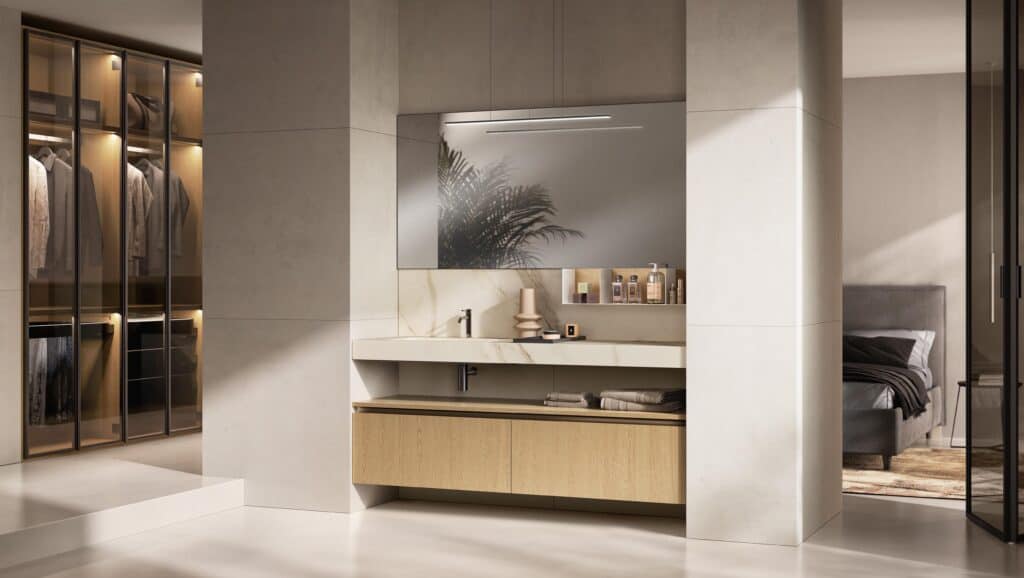 Modern bathroom with a marble sink and wooden cabinetry, adjacent to a walk-in closet with glass doors and a bedroom with a gray bed.