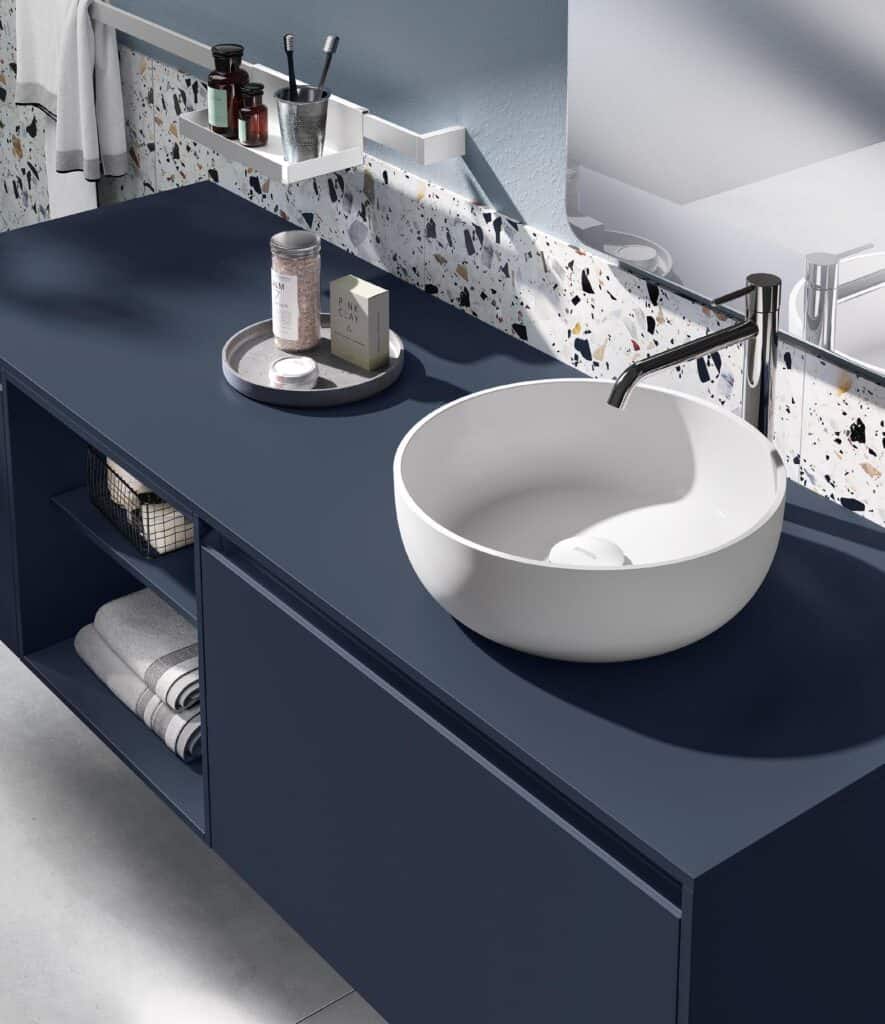 Modern bathroom vanity with a deep blue countertop, a round white vessel sink, a silver faucet, and shelving under the counter. Accessories like a tray with bath items are present, with terrazzo tile backsplash.
