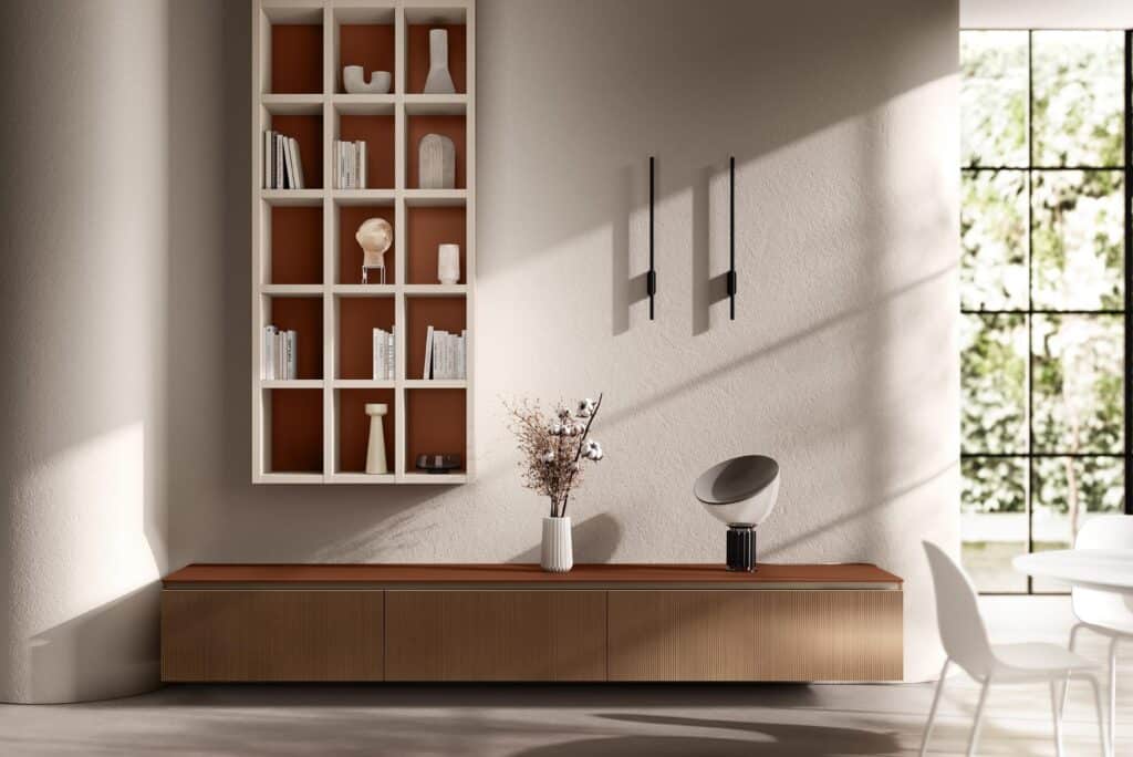 A modern interior with a wall-mounted grid shelf holding books and decorative objects, a long wooden cabinet beneath it, two wall-mounted light fixtures, and a round table with chairs near a large window.