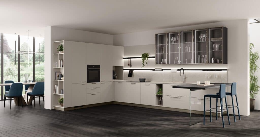 Modern kitchen with white cabinets, a countertop extending to a small bar area with two blue chairs, glass-fronted upper cabinets, and wooden flooring, adjacent to a dining area with blue chairs and large windows showing greenery outside.