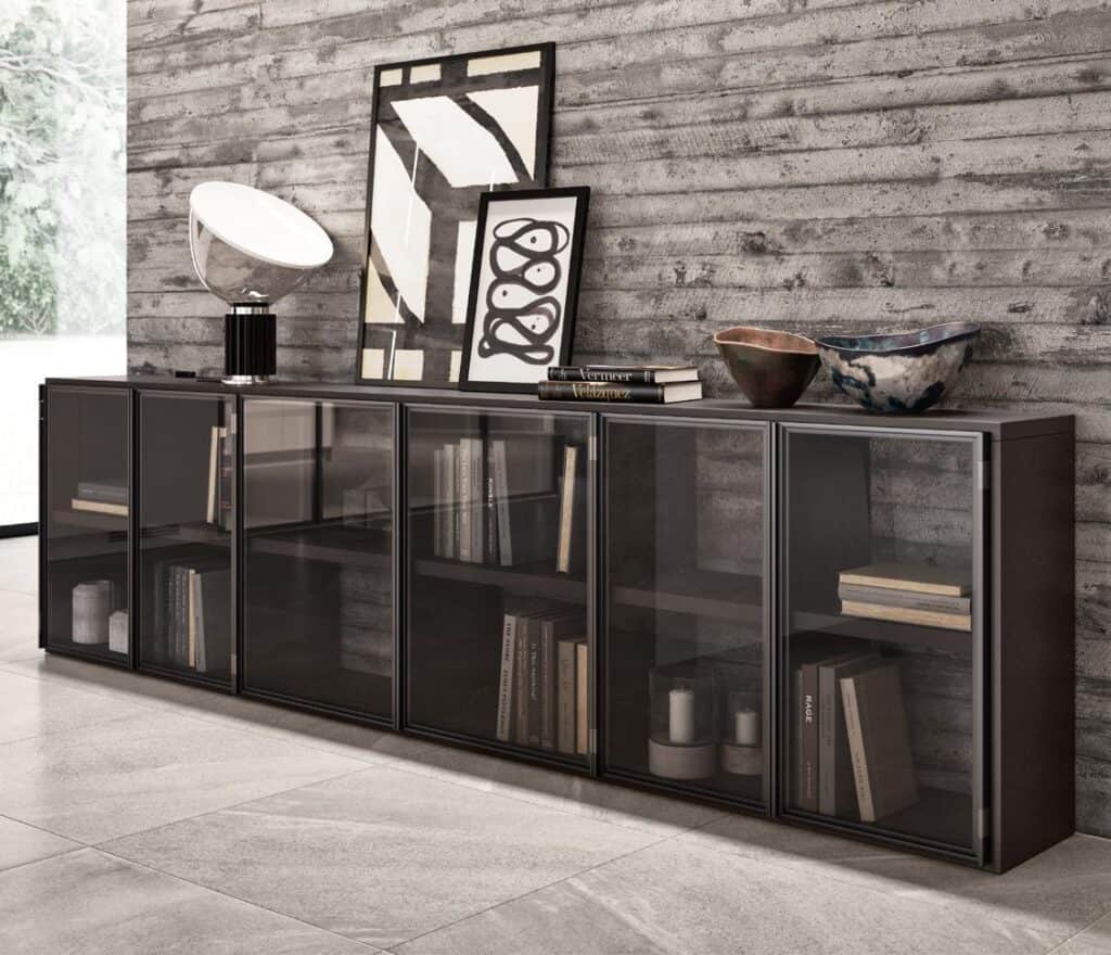 Modern interior with a low black cabinet featuring glass doors, displaying books and decorative items; a textured concrete wall, abstract art, a lamp, and ceramic bowls decorate the top.
