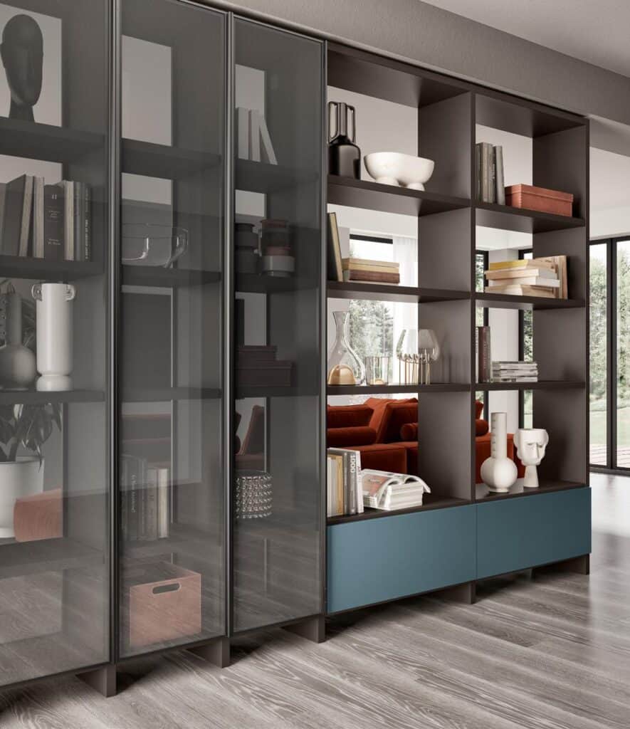 Modern living room with glass-fronted shelves and open shelving unit, decorated with books, vases, and various decor items; visible through the shelves is a red sofa and large windows.