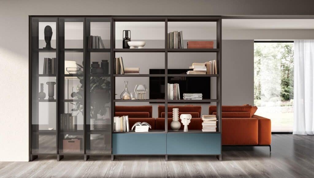 Modern living room with a large bookshelf featuring books, decorative objects, and two blue drawers, a brown sofa, and a view of a garden through a large window.
