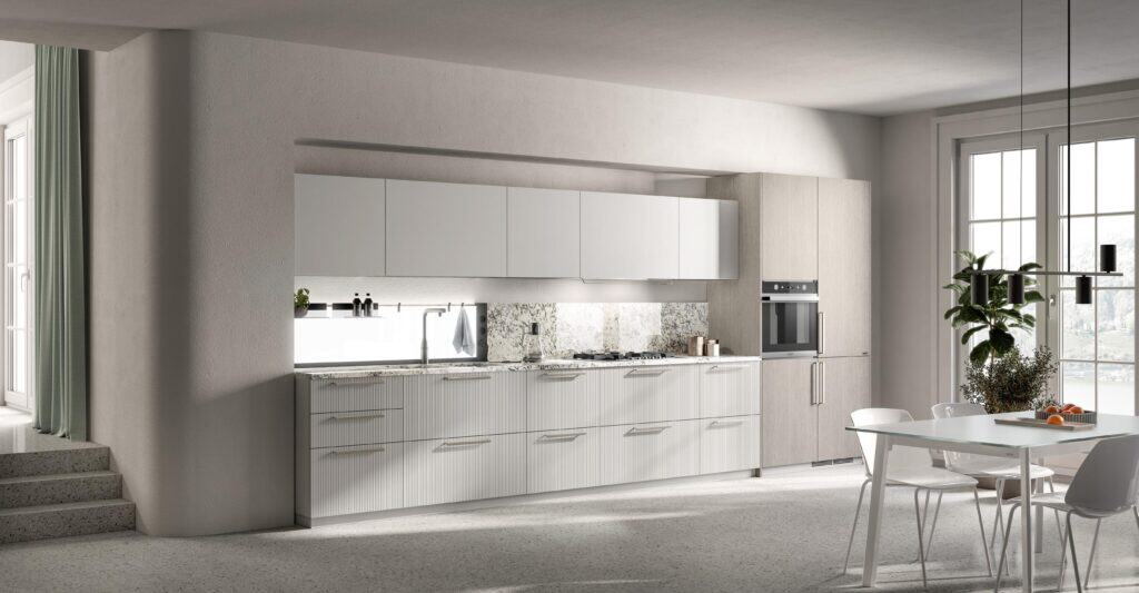 Modern kitchen with white cabinets, marble backsplash, built-in oven, and a sleek dining area with a glass table and white chairs. Pendant lights hang above the table, and large windows provide natural light.