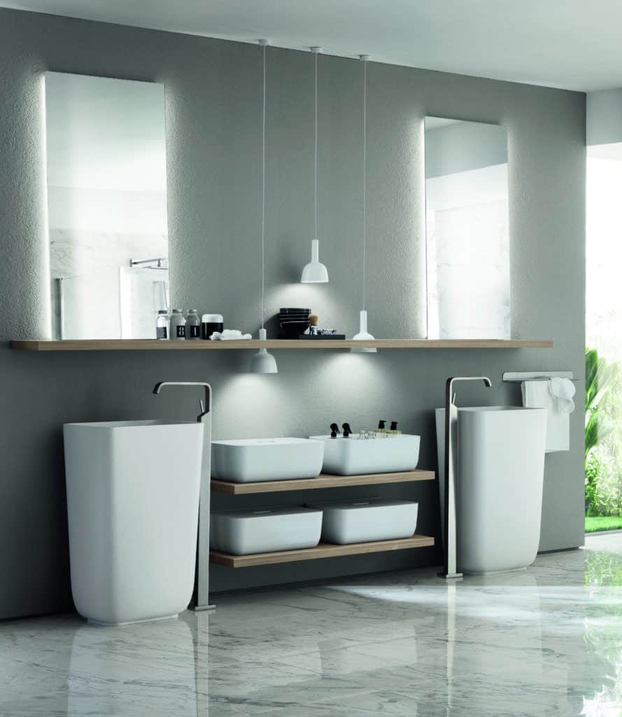 Modern bathroom interior with two tall sinks, a shelf with toiletries, pendant lights, and large mirrors, all set against a gray wall with marble flooring.