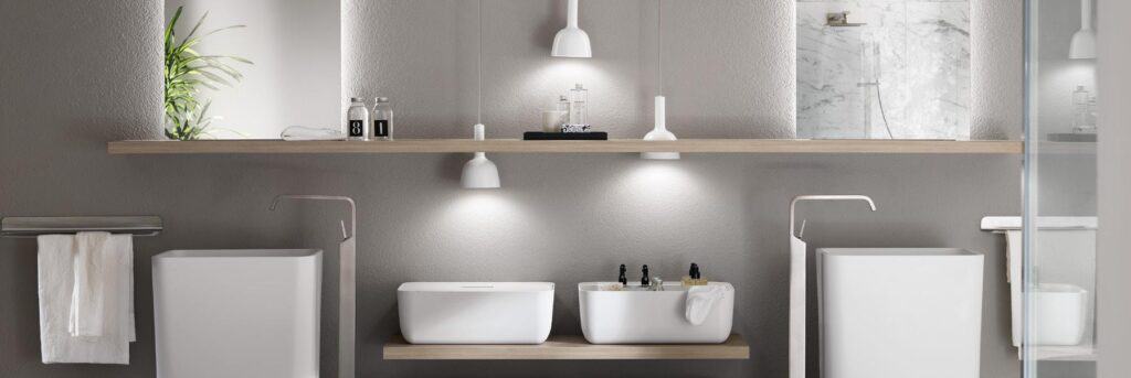 Modern bathroom with two white rectangular basins on a light wood shelf, minimalist fixtures, and soft lighting from two pendant lamps, set against a gray wall.