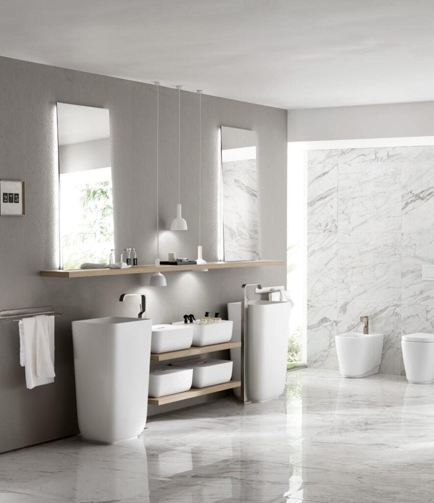 Modern bathroom with two pedestal sinks, a wooden shelf, large mirrors, pendant lights, a towel rack, and marble flooring and walls.