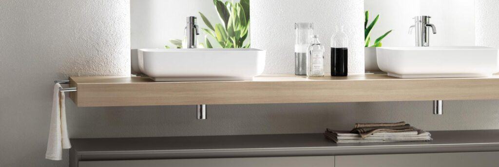 Modern bathroom with two white vessel sinks on a wooden countertop, chrome faucets, decorative bottles, and folded towels beneath.