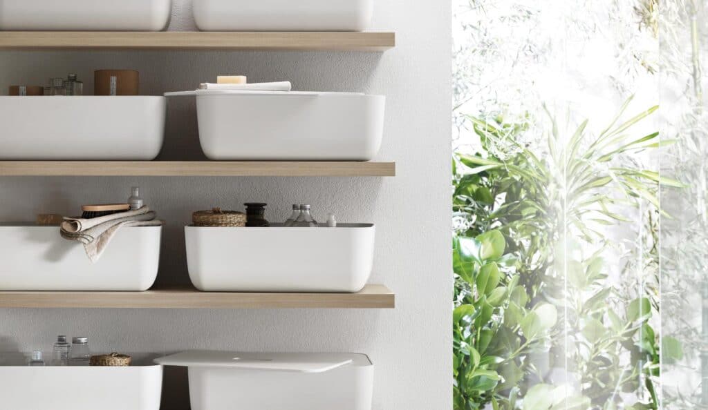 White storage bins on wooden shelves with assorted toiletries, next to a large window with green foliage outside.