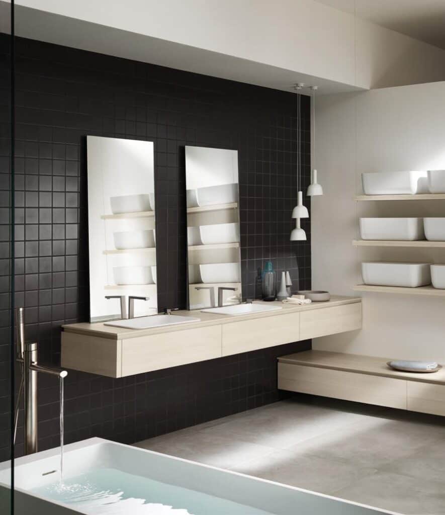Modern bathroom with black tiled wall, dual mirrors and sinks, wooden floating shelves, and a filling bathtub.