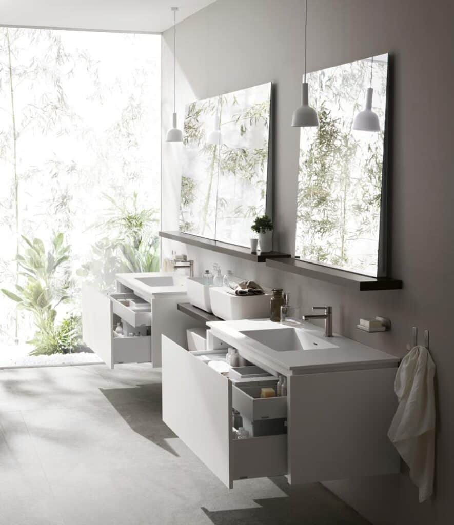 Modern bathroom with minimalist white countertops, open drawers, a large mirror, two pendant lights, and a window view of greenery.