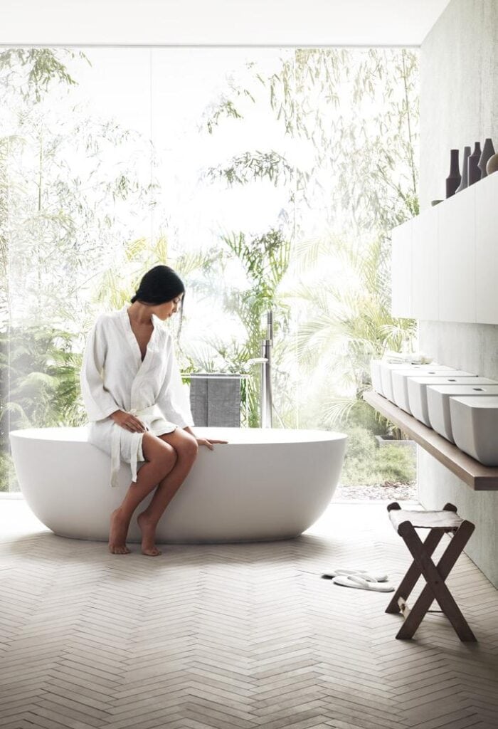 A person in a white robe sitting on the edge of a bathtub in a bright bathroom with large windows and greenery outside.
