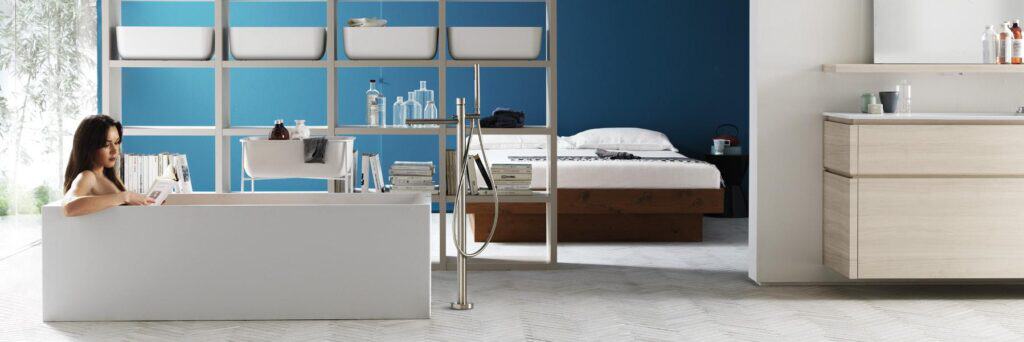 A person is reading in a modern bathroom with an open design, featuring a white bathtub, blue accent wall, and minimalistic decor.