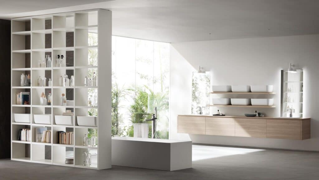 Modern bathroom with a white freestanding bathtub, open shelving filled with books and toiletries, a wall-mounted wooden vanity with rectangular sinks, large windows, and indoor plants.