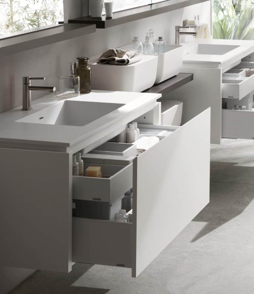Modern bathroom with white vanity, open drawers containing toiletries, and two sinks with chrome faucets.